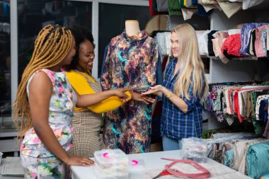A clothing designer shows a dress from her new collection to two interested clients. Two African women and a European woman in the tailors workshop.