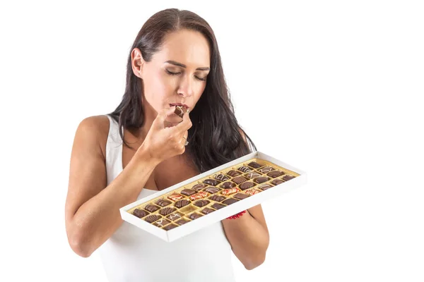 魅力的な暗い髪の女性は フルキャンディーボックスを保持しながら チョコレートキャンディーを食べています 目を閉じてチョコレートがどれだけおいしいかを示しています 隔離された背景 — ストック写真