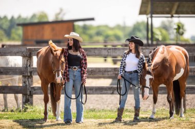 Kahverengi atlı genç kadınlar bir çiftlikte durup yan tarafa bakıp gülümsüyor..