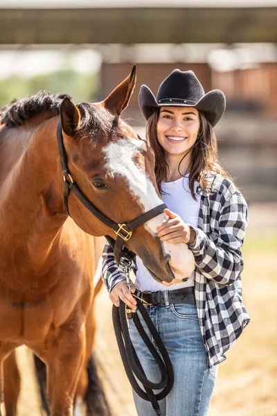 戴牛仔帽的年轻貌美的女人一边在镜头前微笑 一边用手把马打了一下 — 图库照片