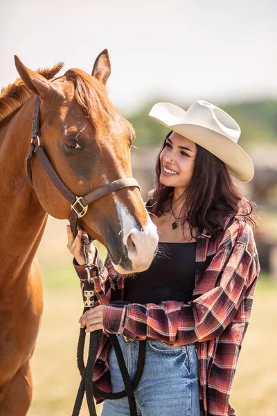 Beautiful girl in a cowgirl wear smiles at her brown horse.