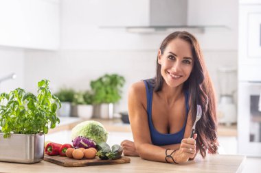 Woman holding a fork leans against the kitchen desk next to assortment of fresh raw vegetables.