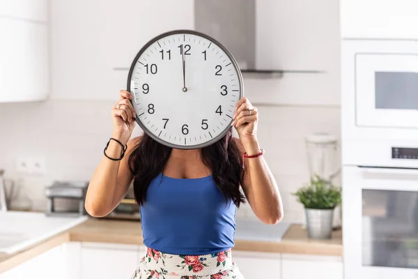 Woman holds a clock showing one minute to twelve in fron of her face.