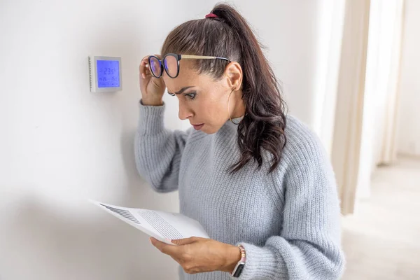 Woman standing next to a thermostat cannot believe the price for heating on her bill due to energy crisis in Europe.