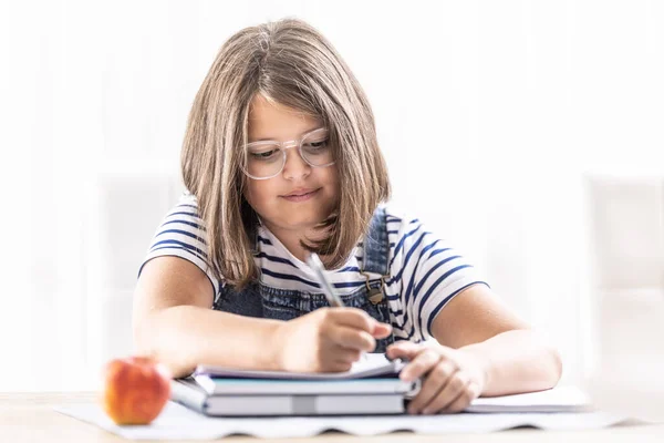 School attending girl wearing glasses writes into an exercise book with a pen having an apple on a table besides her.