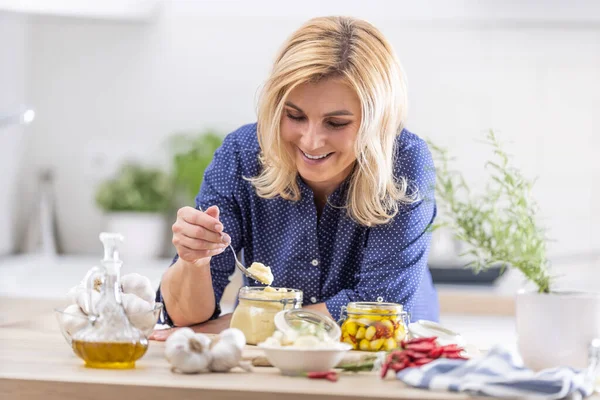 Smiling blonde woman takes spoonful of crushed garlic during preserves making session in the autumn.