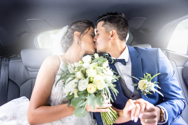 Happy young just married couple kisses in the car holding hands and bride holding a wedding bouquet.