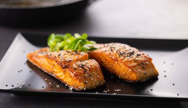 Çıtır çıtır olana kadar pişmiş iki somon fileto, siyah bir tabakta susam serpiştirilmiş..