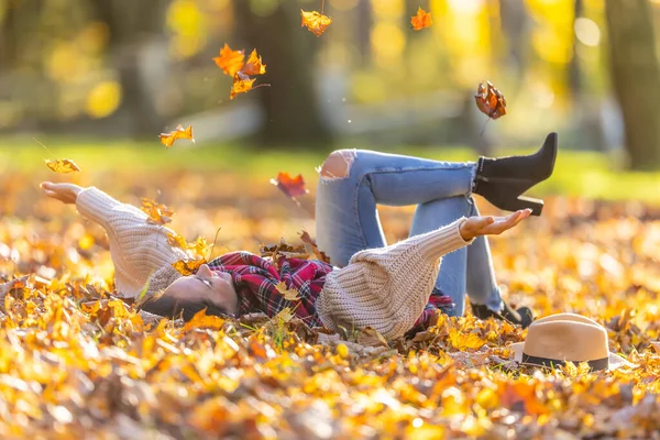 Carefree beautiful brunette lying in the park throws away fallen leaves during sunny autumn sunset.