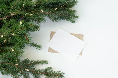 Noel kompozisyonu. Boş tebrik kartı, sahte zarf. Ağaç dalları ve beyaz masa zemininde Noel süslemeleri. Düz, üst manzara.