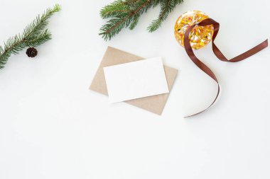 Noel kompozisyonu. Boş tebrik kartı, sahte zarf. Noel cam küresiyle dekorasyon, kahverengi kurdele, köknar ağacı dalları ve çam kozalakları beyaz masa zemininde. Düz, üst manzara