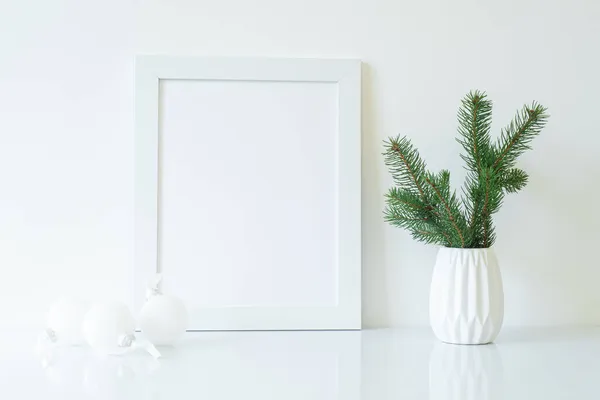 Mock up white frame with fir branch in vase and white christmas decorations on shelf. Empty portrait frame against a white wall