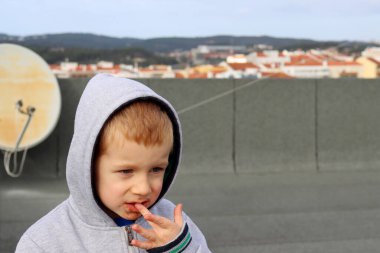A blond boy with a stained chin puts his finger in his mouth. A four-year-old boy in the hood and a dirty face 
