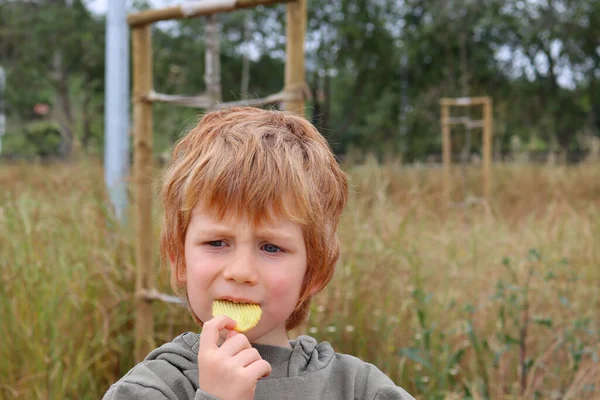 The blond boy is eating chips and looking seriously into the distance. A five-year-old boy eats potatoes with a furrowed brow.