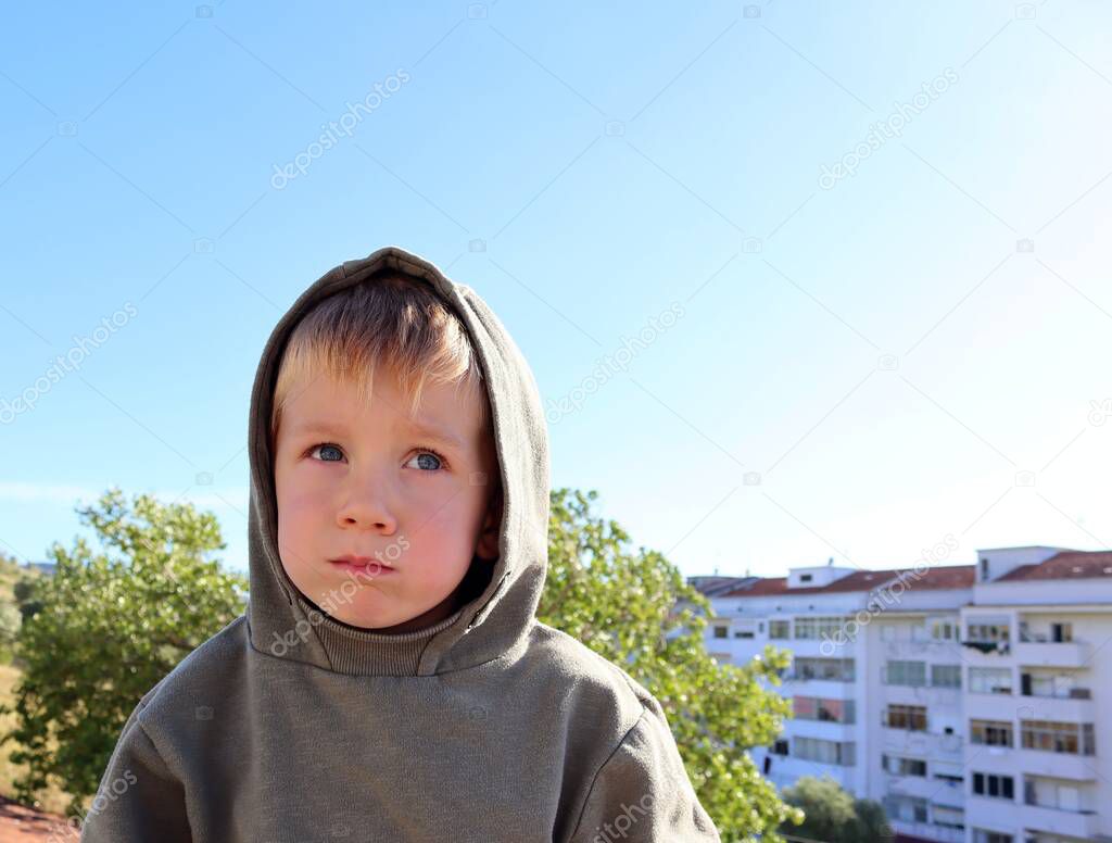 The blond boy in the hood puffed out his cheeks and looks intently into the distance. A thoughtful five-year-old boy. 