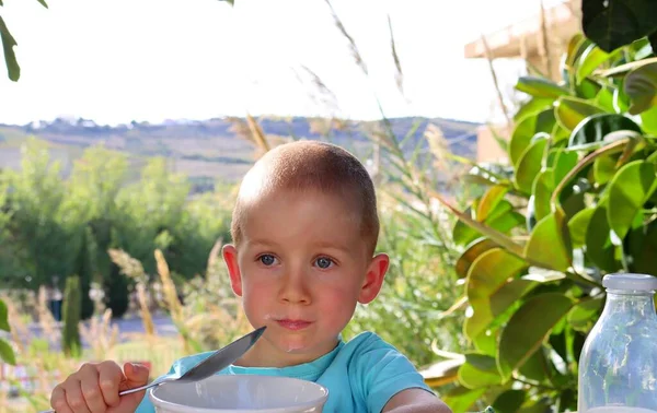 A short-cut, blond boy with a milky mustache stares thoughtfully into the distance. A five-year-old boy eats his lunch outdoors.