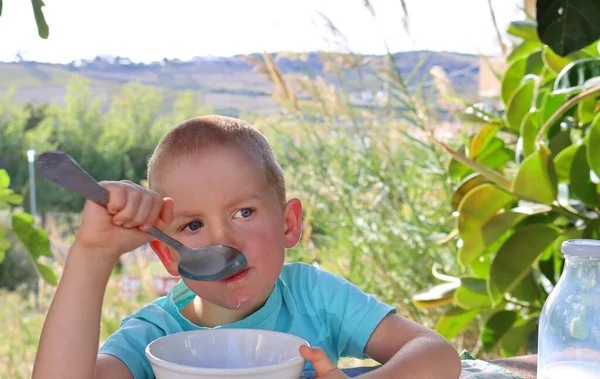 A short-cut, blond boy with a big spoon in his mouth and a thoughtful look. A five-year-old boy immersed in thought.