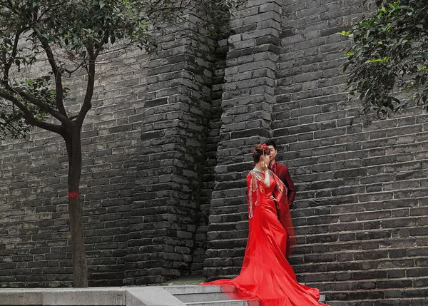 2017年10月22日 古典的な Her と現代の中国の花嫁と新郎 彼の赤いウェディングドレスは 市内の灰色のレンガ造りの壁のふもとに彼らの写真アルバムのポーズ 陝西省 — ストック写真