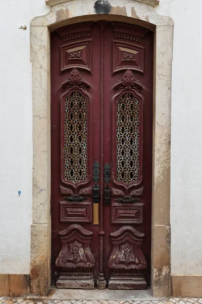 Maroon-brown painted wooden door artistically carved with metal latticework panels-mortar doorframe-heavy decoration of floral motifs-black doorknockers and handles-white wall. Lagos-Algarve-Portugal.