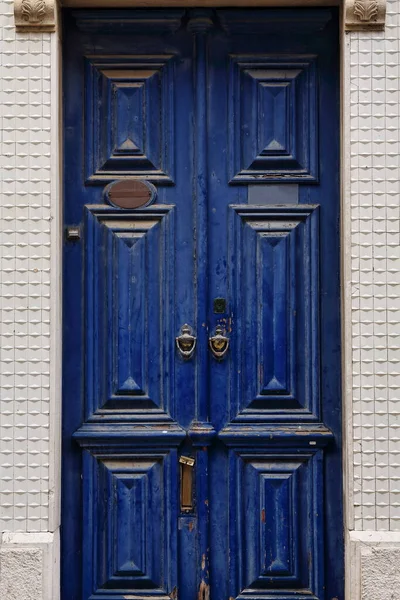 Indigo blue painted wooden door artistically carved with relief panels-vertical letterbox slot-old metal doorknockers and handles-chipped paint-floral mortar lintel-white wall. Lagos-Algarve-Portugal.