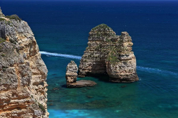 Eastwards view from atop the sheer cliff sheltering Praia dos Pinheiros Beach to the Leixao da Balanca-Scales Sea Stack facing the sandy spot, white wake of bygone motorboat. Lagos-Algarve-Portugal.