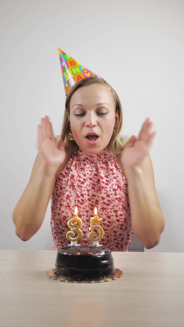 Celebración de cumpleaños en línea. Webcam retrato de una joven feliz con una gorra de fiesta, soplando una vela en un pastel, soplando un silbato, disfrutando de unas vacaciones en casa, cámara lenta. vídeo vertical — Vídeo de stock