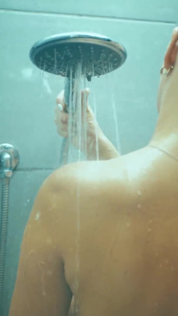 Rear view close up of a young Caucasian woman with long hair standing under the shower in a bathroom, washing and rinsing her hair, slow motion — стоковое видео