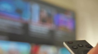 Akıllı TV izleyen ve modern siyah uzaktan kumanda kullanan adam, arka planda bulanık televizyon. Görüntüyü Kapat - DCi 4K Video