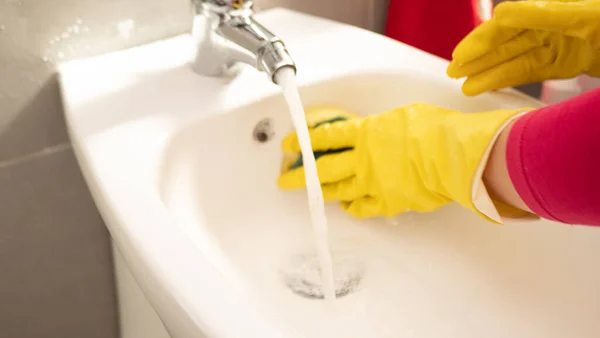 打扫你的房子。妇女在浴室做家务活，手戴黄色手套清洁水龙头，钢制洗涤槽，带有蓝色抹布和洗涤剂喷雾 — 图库照片