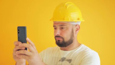 Sarı arka plandaki inşaat işçisi telefonla fotoğraf çekiyor.
