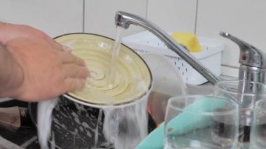 Bir adam, modern bir mutfakta kirli bulaşıkları musluğun altında yıkıyor.
