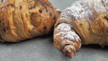 Gri bir arka planda iki taze kruvasan, bir çikolatalı kruvasan ve beyaz parçalı bir kruvasan..