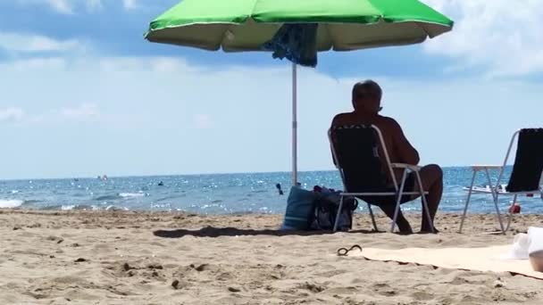 Een Man Zit Onder Een Paraplu Het Strand — Stockvideo
