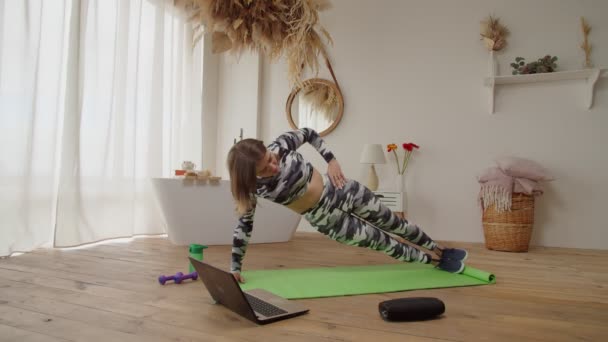 Behoorlijk actieve sportieve fit vrouw doen side plank oefening na online yoga klasse binnen — Stockvideo
