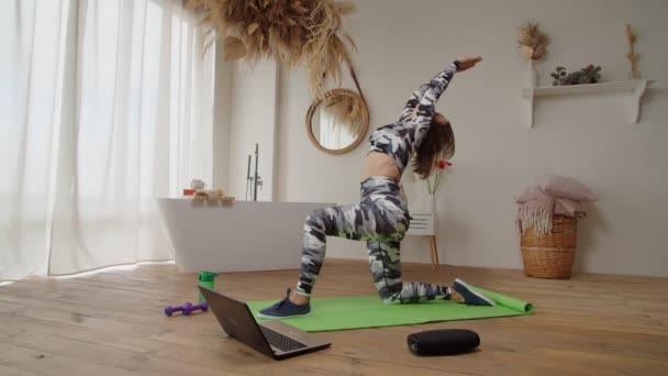 Jolie sportive femme fitness avec ordinateur portable sur tapis d'exercice faisant croissant de lune pose à l'intérieur — Video