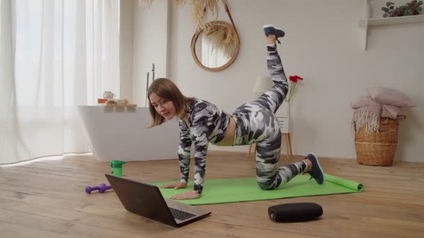 Güzel, sportif formda bir kadın dizüstü bilgisayarda eğitim videosu izliyor, evde eşek tekmesi yogası yapıyor. — Stok video