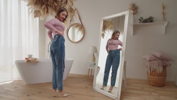 Довольно счастливая женщина демонстрирует свою потерю веса, надевая старые джинсы в помещении — стоковое видео