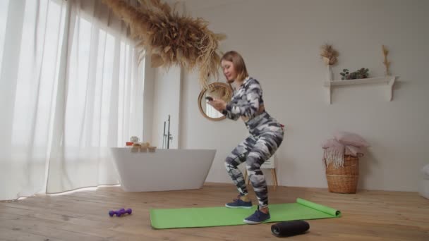 Прекрасна спортивна жінка, що переглядає онлайн на мобільному телефоні і робить присідання вправи вдома — стокове відео
