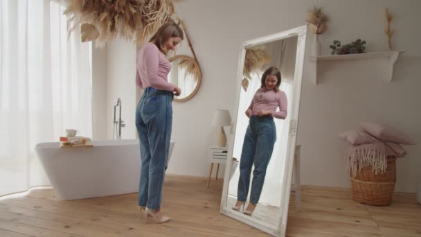 Веселая стройная взрослая женщина в джинсах, проверяющая в зеркале свой успех потери веса дома — стоковое видео