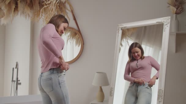 Malheureuse jolie femme essayant de serrer dans une vieille paire serrée de jeans devant le miroir — Video