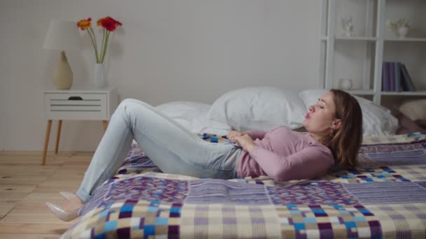 Mujer bonita trastornada acostada en la cama, luchando para encajar en jeans ajustados después de ganar peso en interiores — Vídeos de Stock