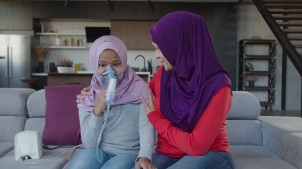 Madre musulmana positiva apoyando a la pequeña hija enferma haciendo tratamiento del asma con nebulizador — Vídeos de Stock