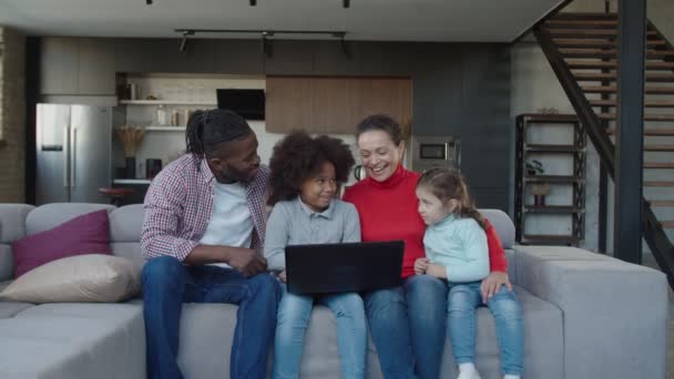 Весела багатоетнічна сім'я з двома милими змішаними расовими дочками, що спілкуються онлайн на ноутбуці в приміщенні — стокове відео