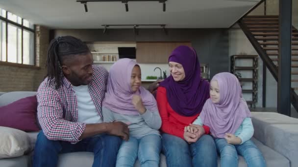 Boldog multiracionális muszlim család kislányokkal hidzsábos kötődésben és a kanapén való osztozkodásban. — Stock videók