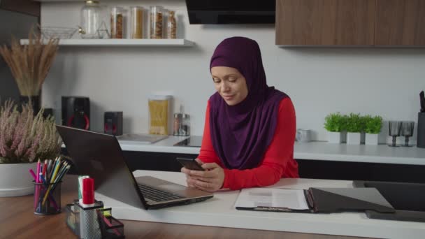 Mooie moderne moslim vrouw in hijab tekstberichten online op smartphone in de huiselijke keuken — Stockvideo