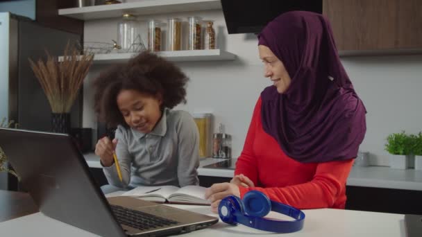 Opiekuńcza muzułmańska matka pomaga wielorasowej córce w wieku szkolnym w odrabianiu lekcji w domu — Wideo stockowe