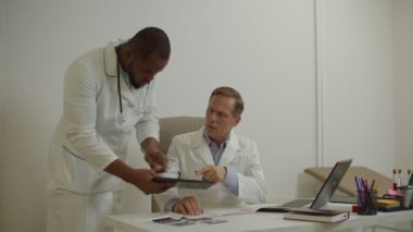 Afrikalı Amerikalı doktor ve deneyimli erkek meslektaşı tablet bilgisayardaki tıbbi test raporunu tartışıyorlar.