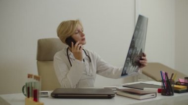 Orta yaşlı, sevimli bir doktor MRI taramasına bakıyor ve tıp bürosunda cep telefonuyla konuşuyor.