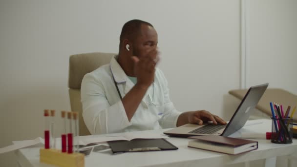 Allegro medico maschio nero in cuffie mani libere godendo la musica mentre si lavora sul computer portatile — Video Stock