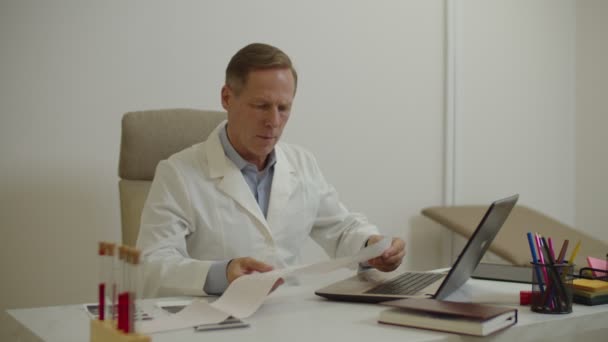 Zuverlässiger männlicher Arzt untersucht Elektrokardiogramm des Patienten und tippt EKG-Testergebnis auf Laptop — Stockvideo